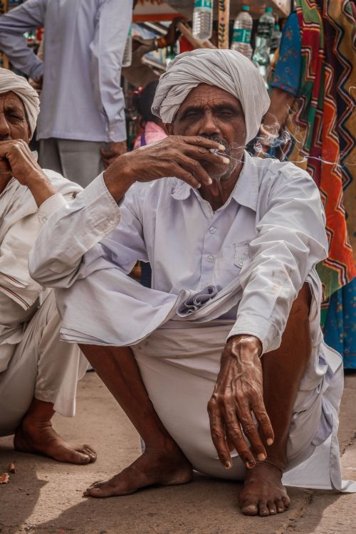 Free Elderly man smoking, dressed in traditional clothing, crouching outdoors in Asia. Stock Photo