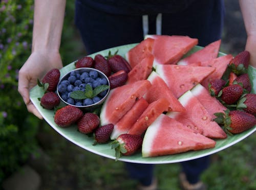 무료 얇게 썬 수박, 딸기, 블루 베리가 가득한 타원형 녹색 접시를 들고있는 사람 스톡 사진