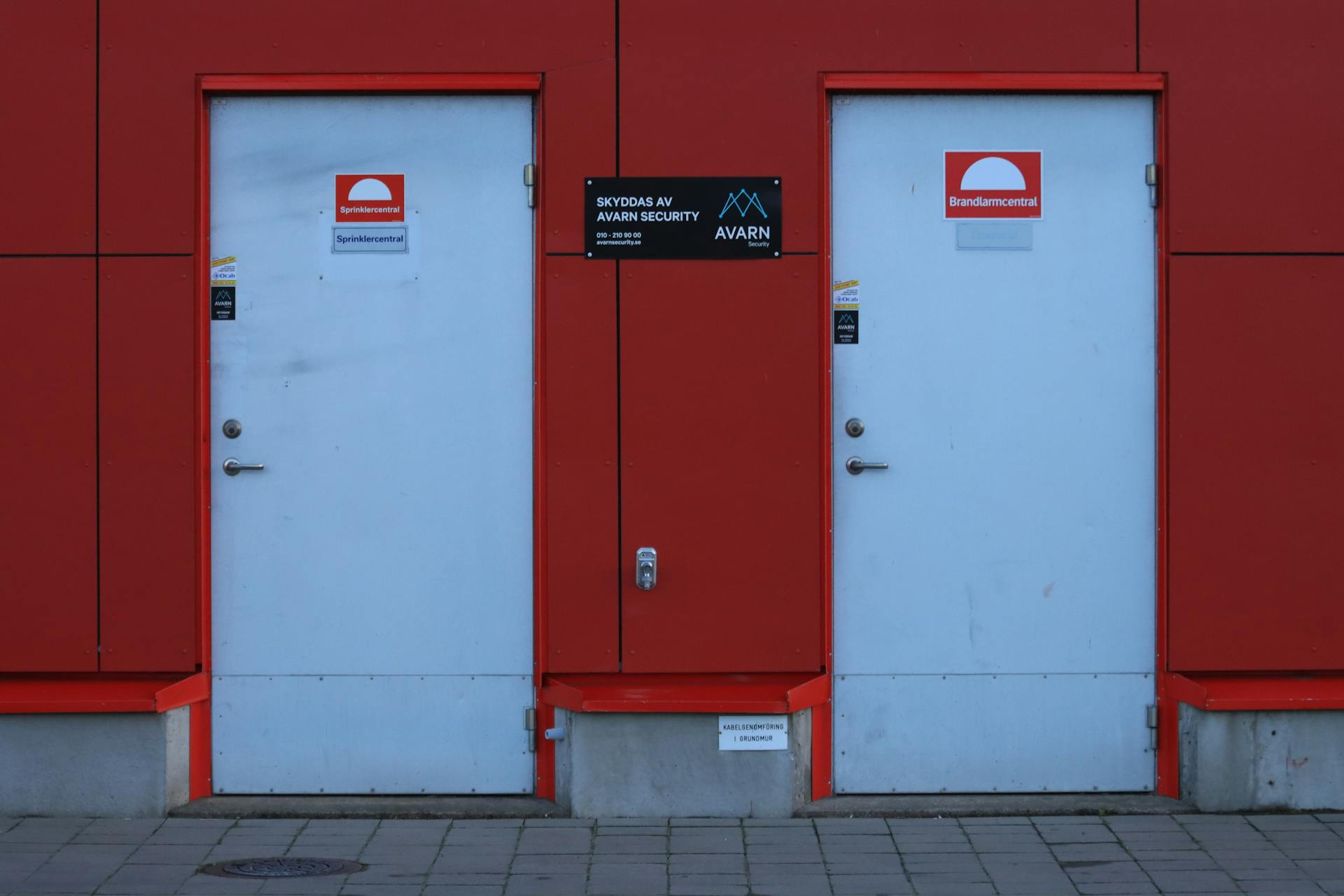 Two white doors against a red wall featuring security signs and logos.