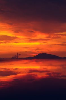 Breathtaking sunset with vibrant tones reflecting on water, highlighting silhouettes of mountains and industrial skyline.