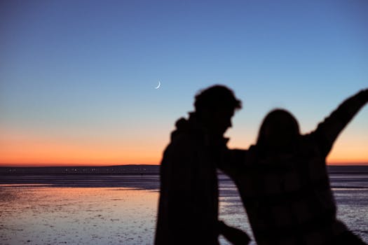 Silhouetted couple enjoying the serene beach twilight with a crescent moon in the sky.