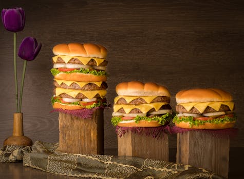Three stacked cheeseburgers with wooden backdrop and tulip decor, emphasizing fast food indulgence.