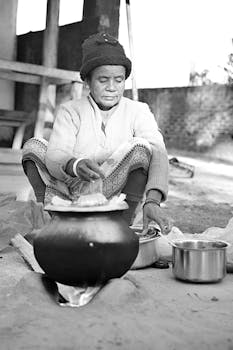 A senior woman prepares food outdoors in black and white with traditional cookware.