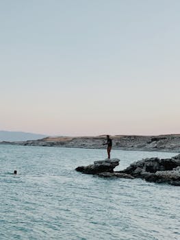 A man stands on a rock overlooking the sea in Qurago, Iraq, with mountains in the background.
