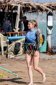Smiling woman wearing a life vest holding a kiteboard, enjoying a summer day at the beach.