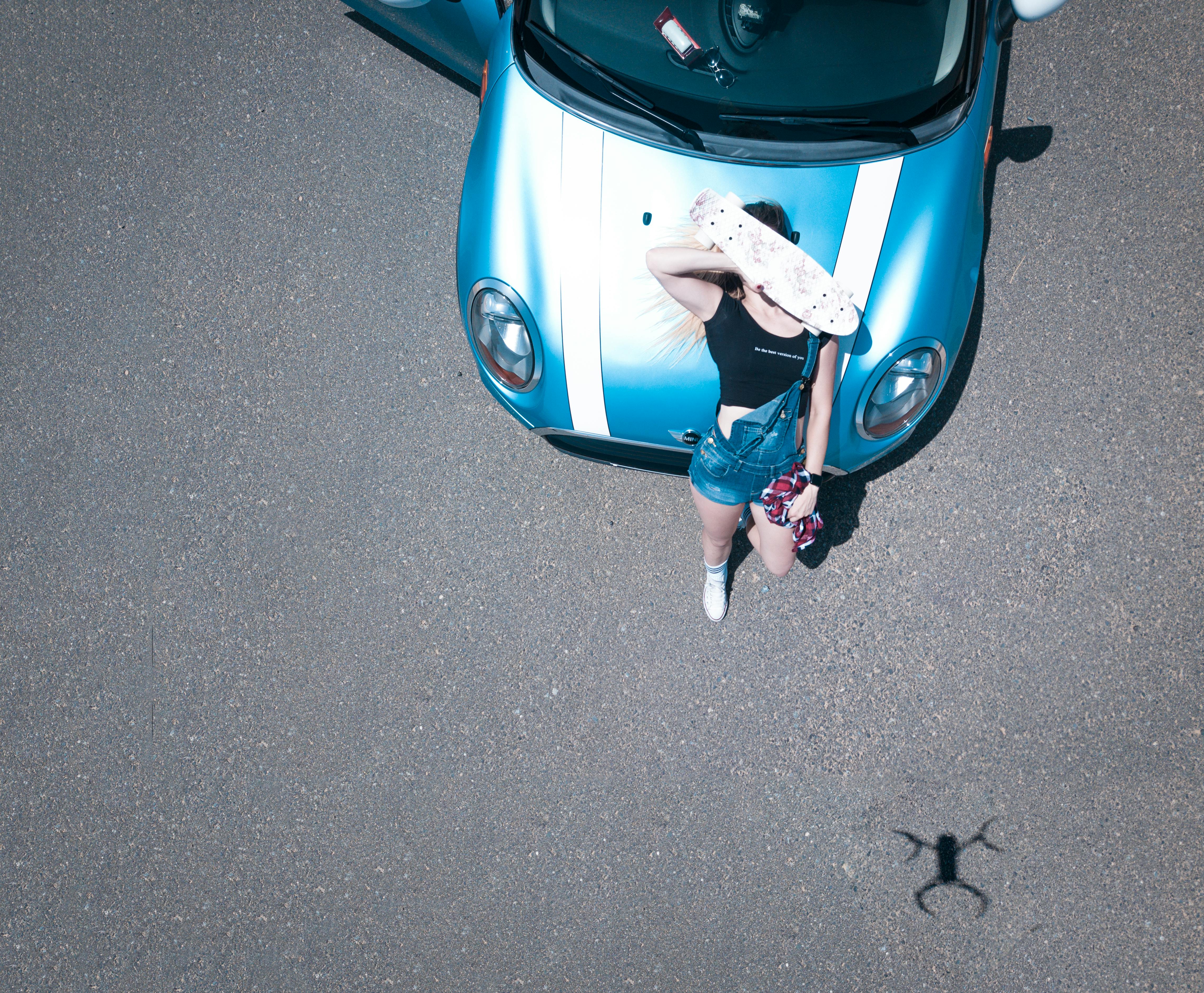 无人机画面中，一名女子正靠在蓝色汽车上，阳光明媚，她手里还拿着滑板