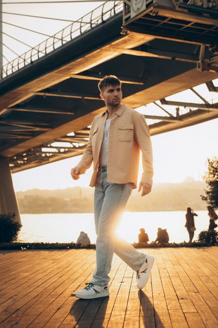 Young man in fashionable attire walking on a boardwalk in Turkey, with a bridge and sunset in the background.