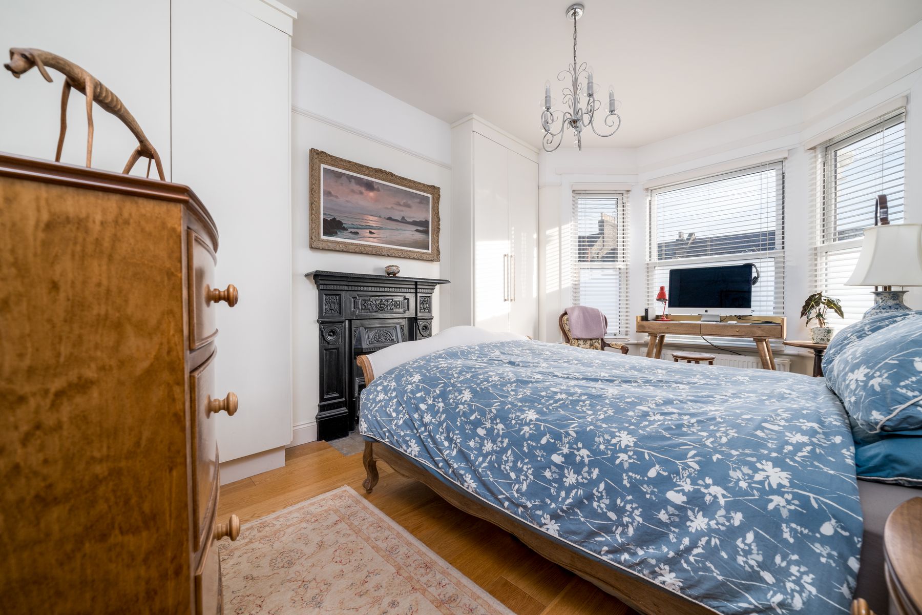 Bedroom with blue floral bedding, old wood furniture, black fireplace, small desk, chandelier and bay windows
