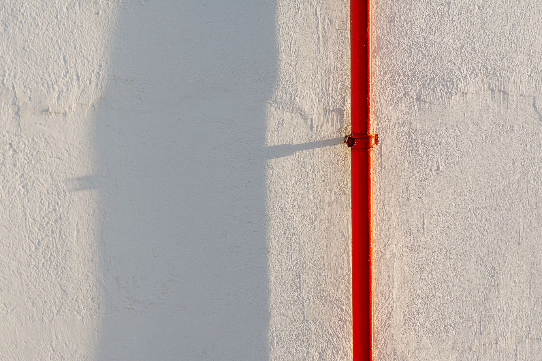 A striking red pipe casts a shadow on a textured white concrete wall.