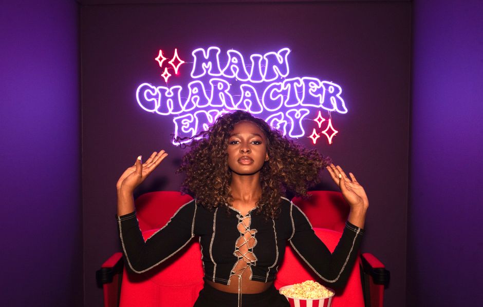 Woman with curly hair posing confidently in front of neon sign and popcorn.