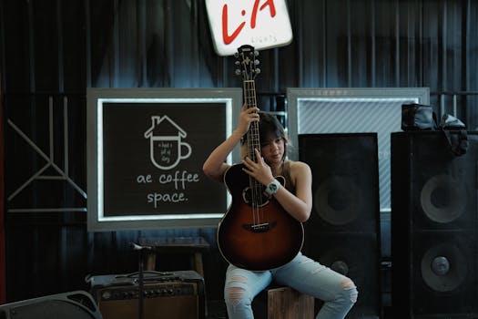 A young woman sits on stage with a guitar in a coffee shop ambiance, featuring signage and musical equipment.