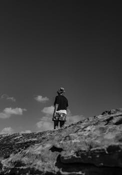 Black and white photo of a solitary hiker standing on a rocky outcrop under open sky.