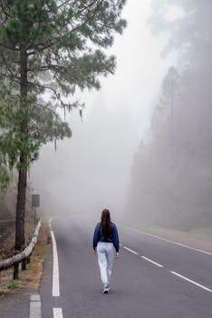 A woman walks alone on a misty countryside road surrounded by foggy trees, evoking solitude and adventure.