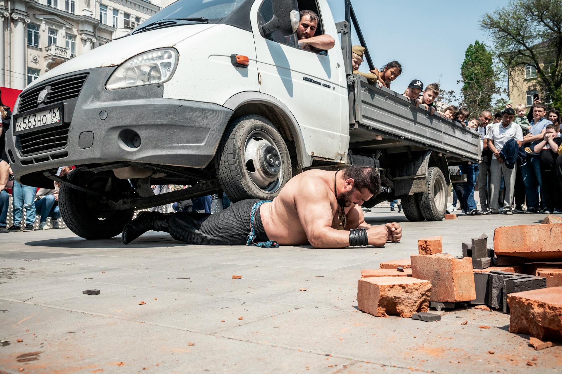 A strongman performs a stunning feat by lifting a truck while a crowd watches outdoors.