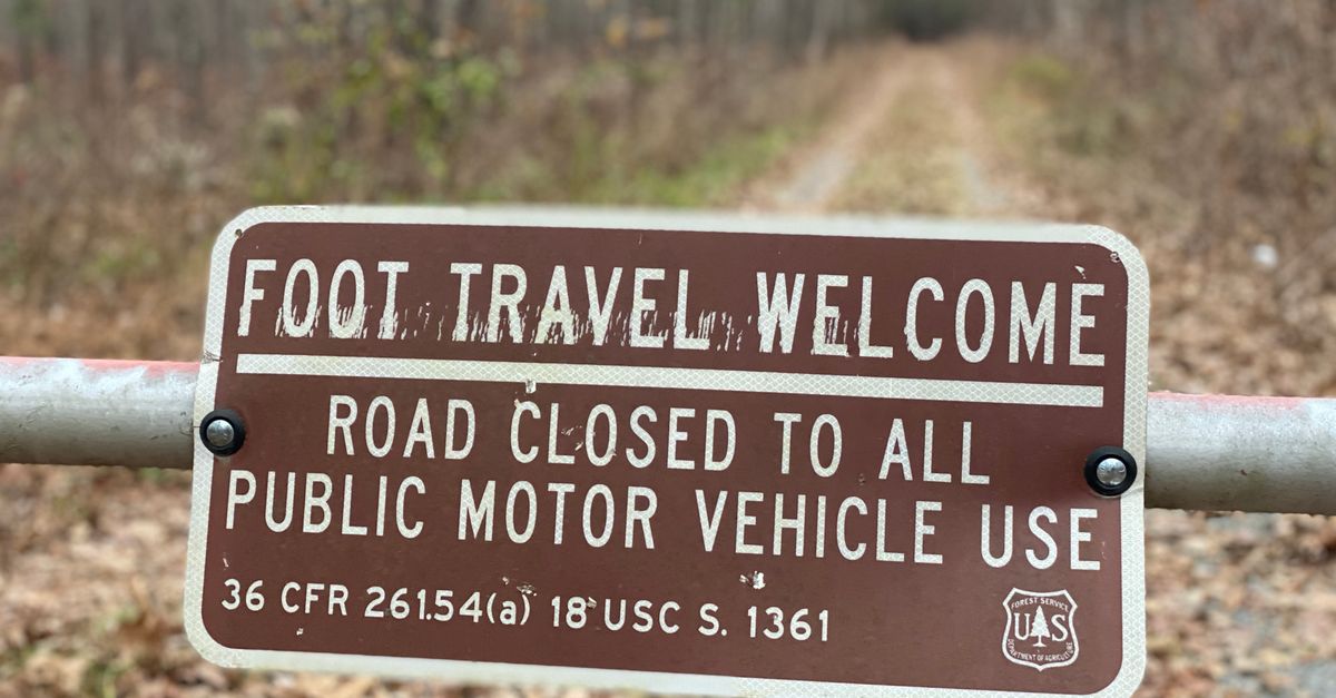 A sign in a forest indicating road closed to vehicles, welcoming foot travel only.