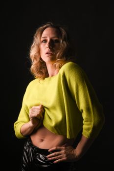 Stylish blonde woman in a neon sweater poses confidently against a dark studio backdrop.