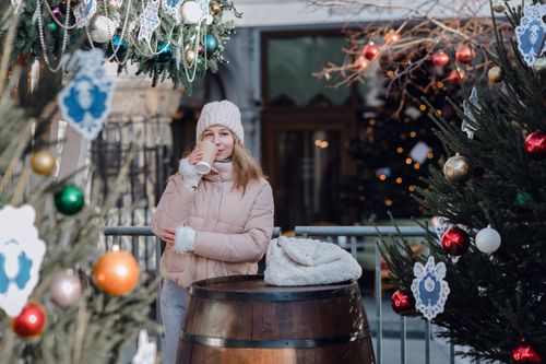 Free A woman in winter attire sips a hot drink at a festive outdoor market with Christmas decorations. Stock Photo