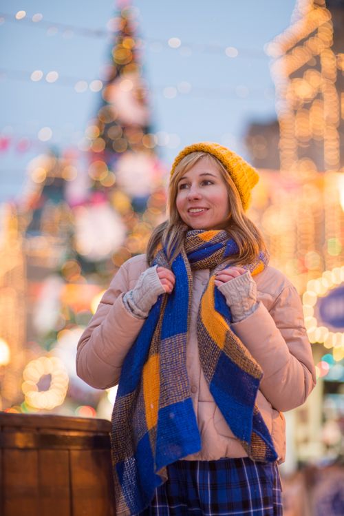Free Woman in winter attire smiling at a colorful Christmas market filled with lights. Stock Photo