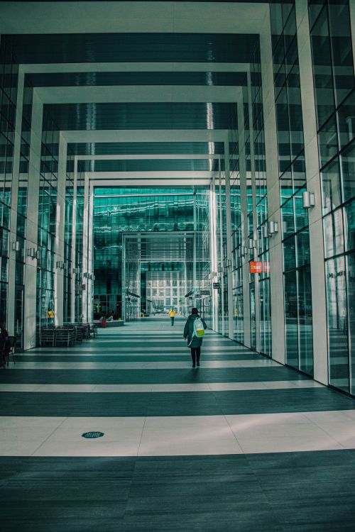 Free A person walks through a geometrical modern glass corridor in an office building, featuring reflections and turquoise tones. Stock Photo