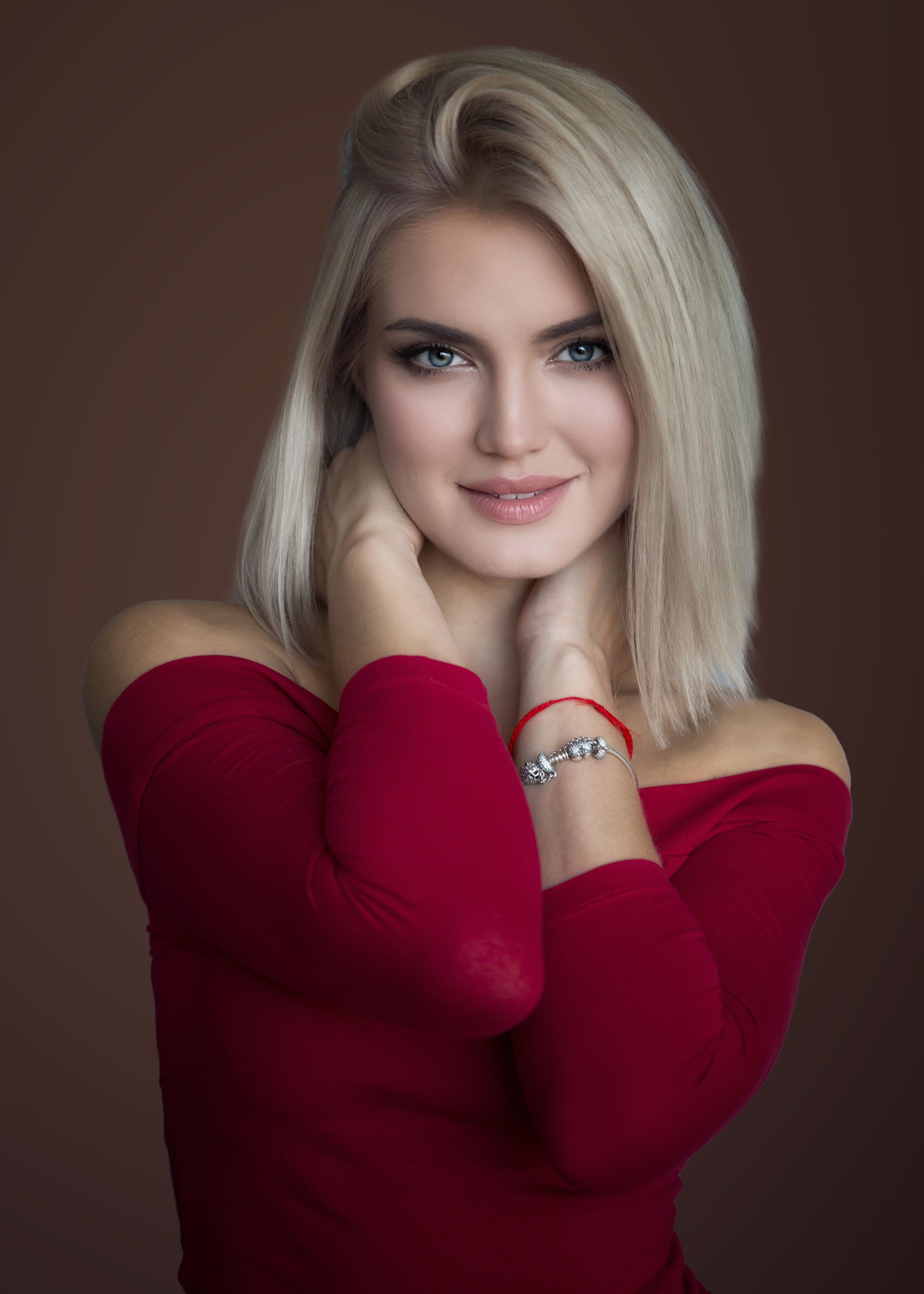 Studio portrait of a blonde woman wearing a red blouse, smiling softly with jewelry accessories.