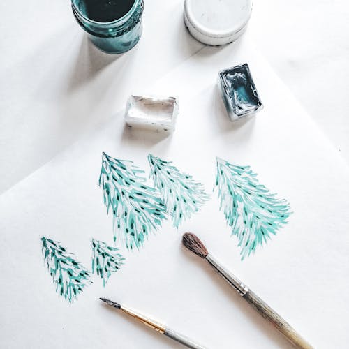 Top View of Paintings of Christmas Trees on a Paper next to Paint Bottles and Paintbrushes 