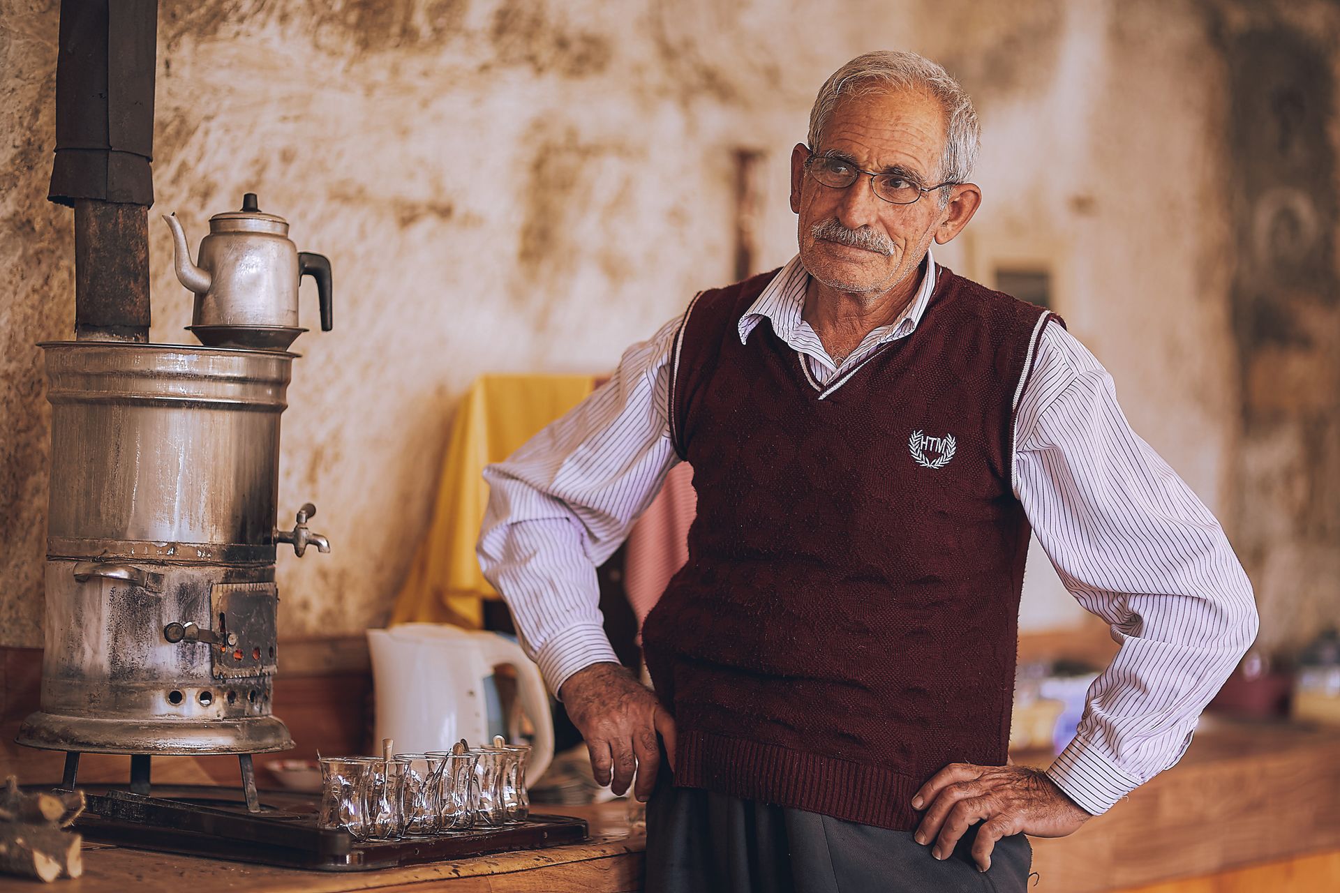Senior man in glasses stands by a traditional furnace in a rustic kitchen.