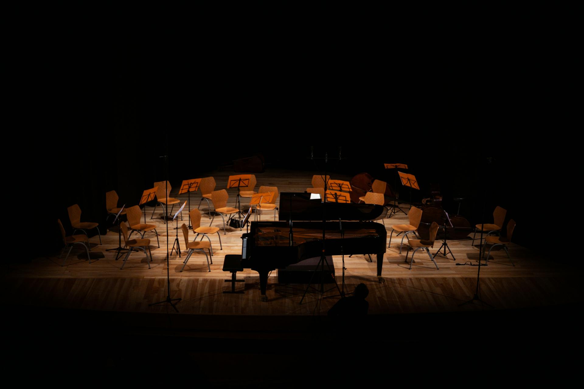 A dimly lit concert hall stage featuring an empty grand piano and chairs for an orchestra.