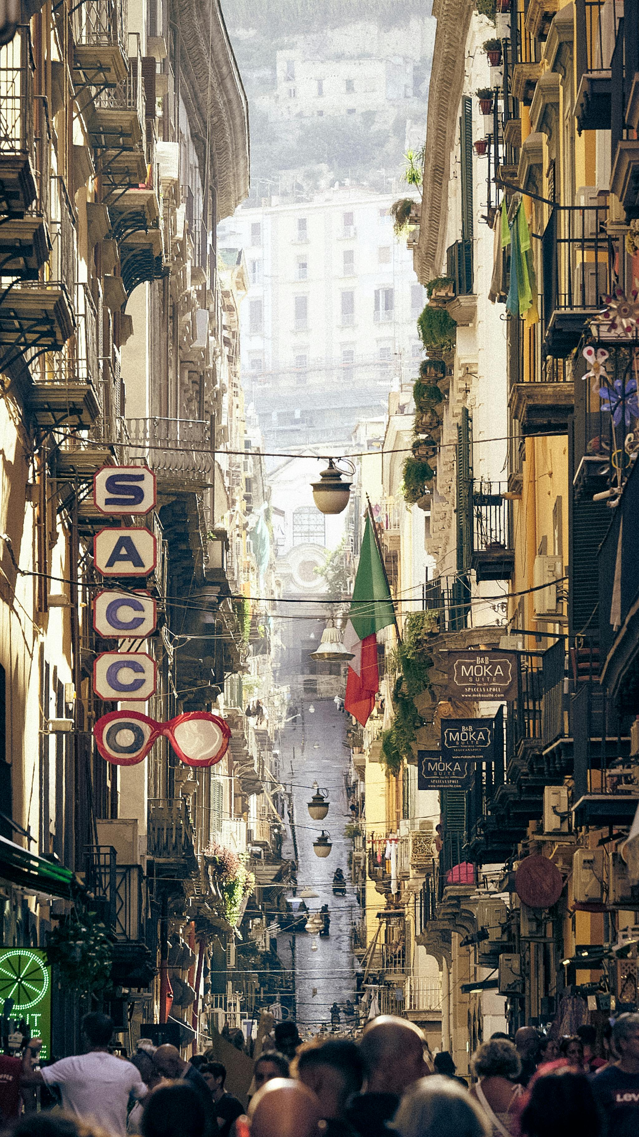 Bustling narrow street in Naples with vibrant buildings and Italian flags, capturing the essence of urban life.