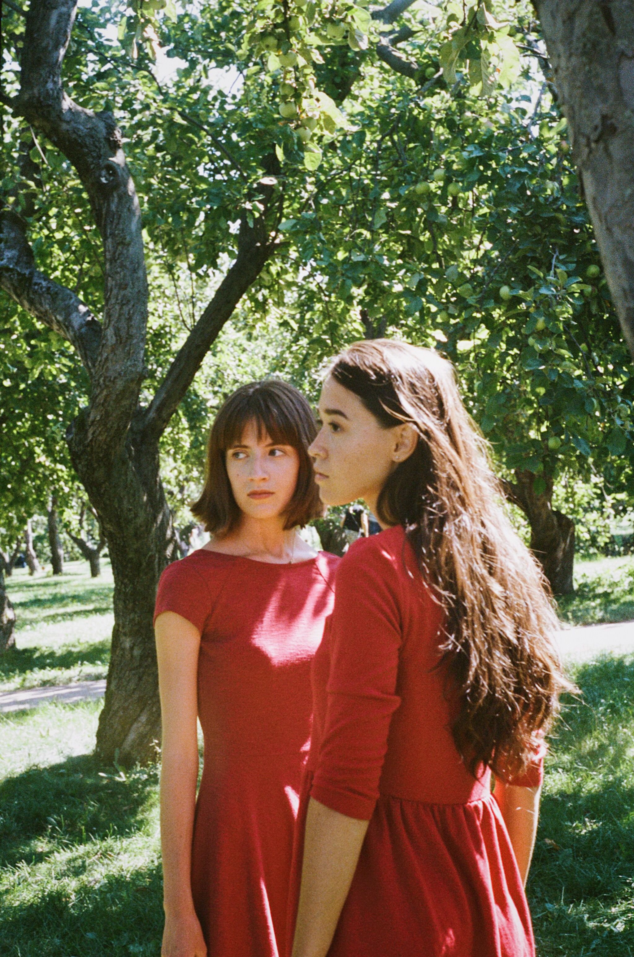 Two women in red dresses enjoy a sunny day in a lush green park.