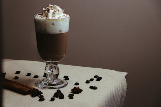 A stylish glass of coffee topped with whipped cream and surrounded by coffee beans and cinnamon sticks.