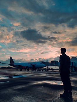 A man silhouetted against a vibrant sunset sky while observing a parked airplane at the airport.