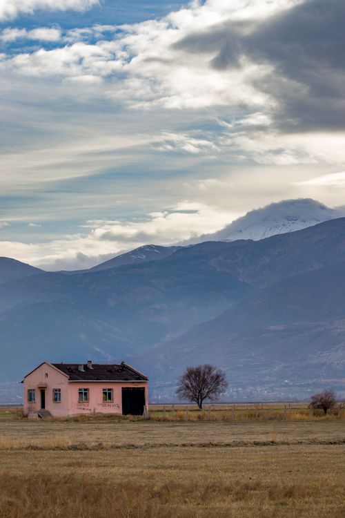 Free An abandoned rural house in a field with a mountain backdrop under a cloudy sky. Stock Photo