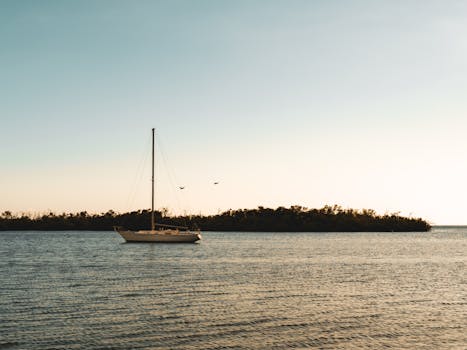 A serene image of a sailboat at sea during sunset, with a clear sky and distant coastline.
