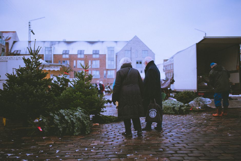 An elderly couple explores a festive outdoor Christmas market with trees and holiday decorations.
