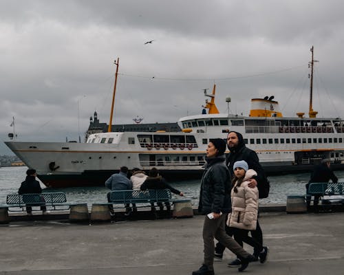 A Family Wearing Winter Jackets Walking Near Ship on Body of Water
