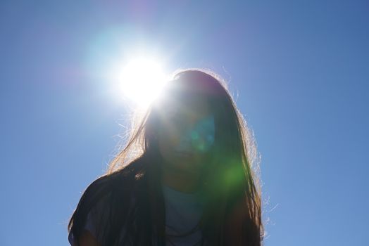 Backlit portrait of a woman with sun flare against a bright blue sky.