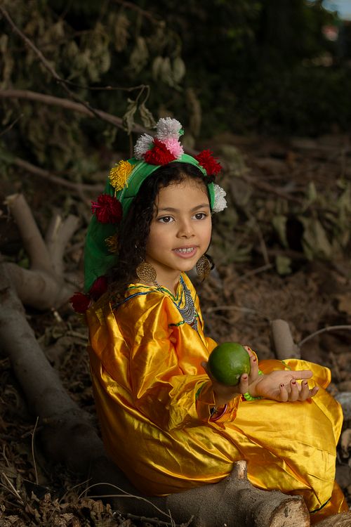 Free A young girl in vivid cultural clothing holds fruit, sitting outdoors, radiating innocence and joy. Stock Photo