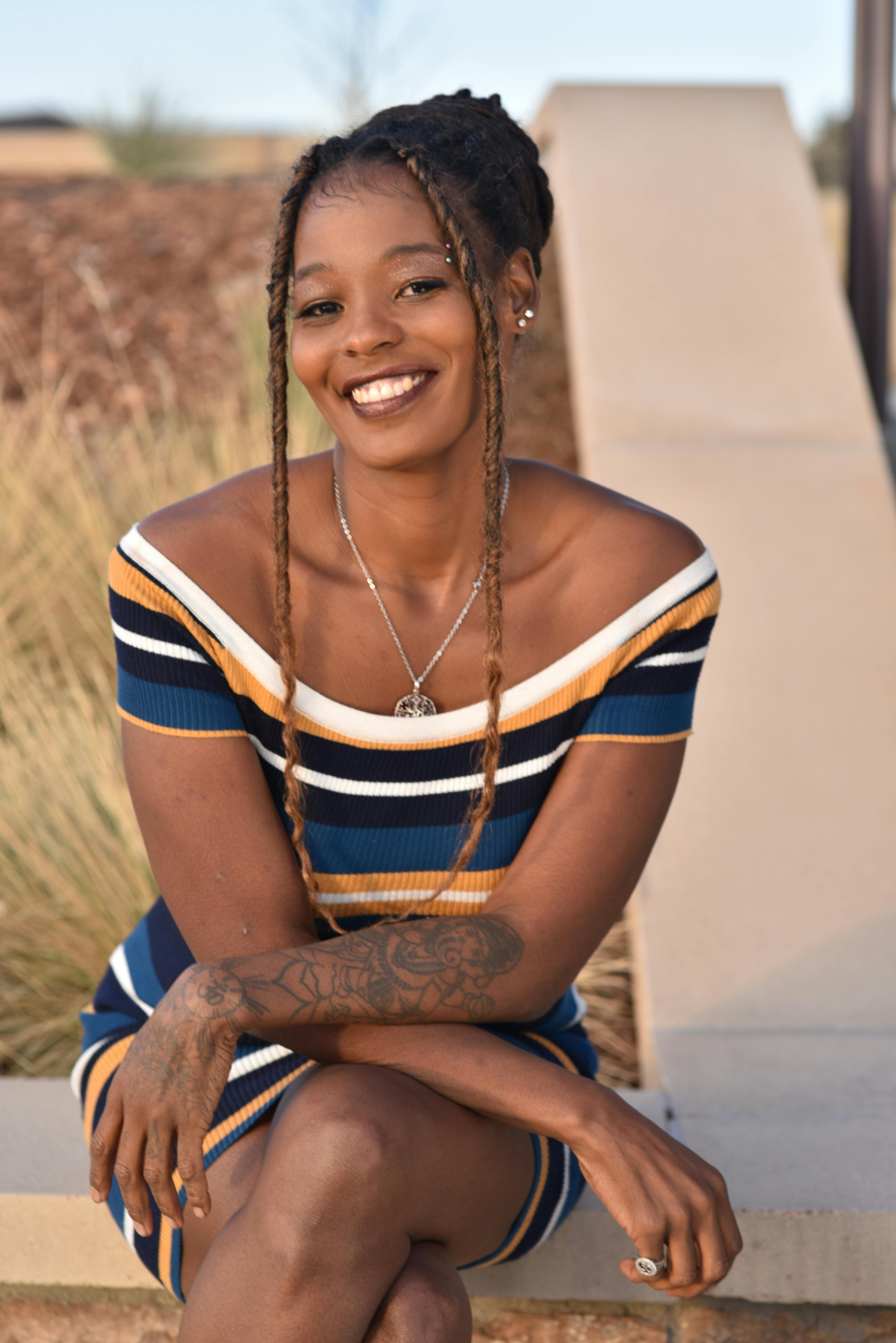 African American woman smiling and posing with crossed legs outdoors in a striped dress.