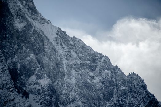 Dramatic view of a snow-covered rocky mountain peak under a cloudy blue sky.