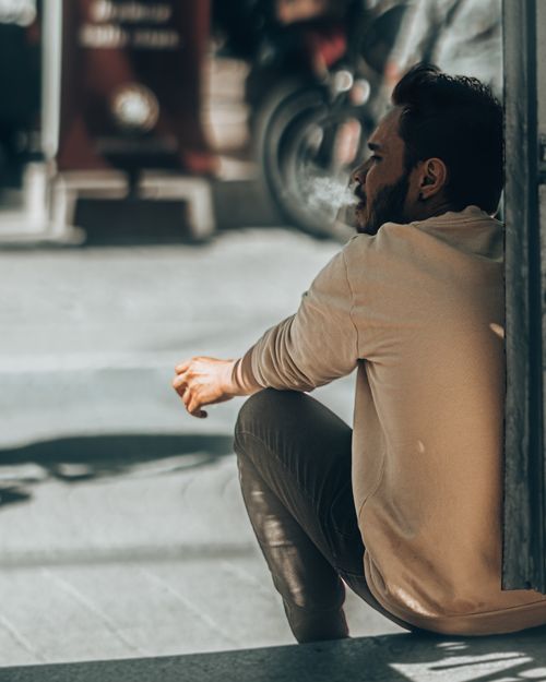 Free A contemplative bearded man sits on the street, casting a shadow in a sunlit urban environment. Stock Photo