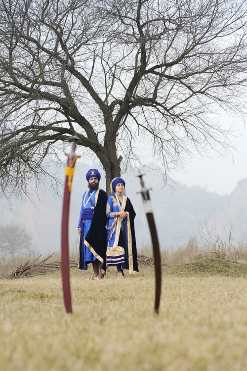 Free A couple in traditional garments standing by a tree with swords in a grassy landscape. Stock Photo
