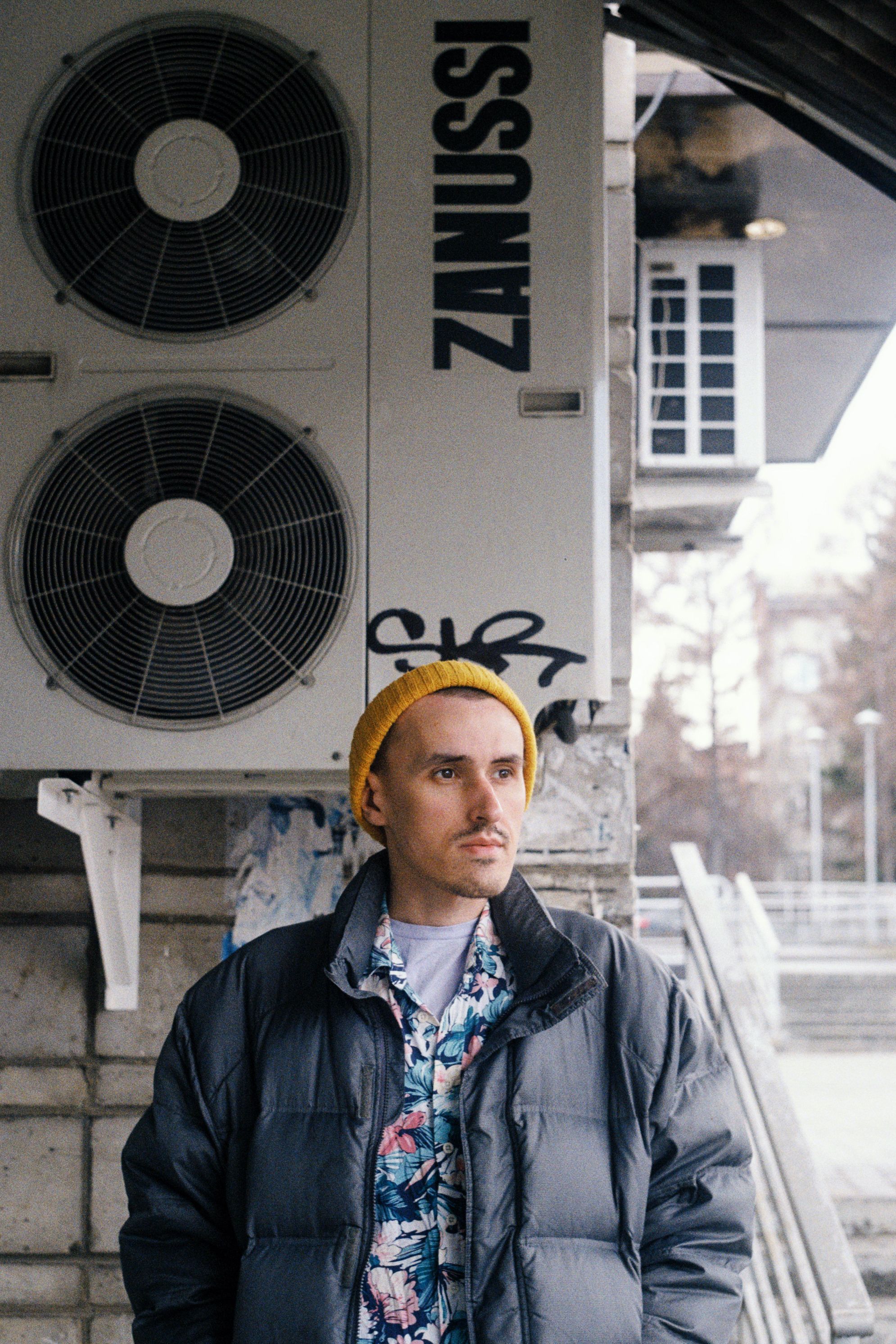 Stylish man in warm jacket and beanie standing by industrial backdrop, urban setting.