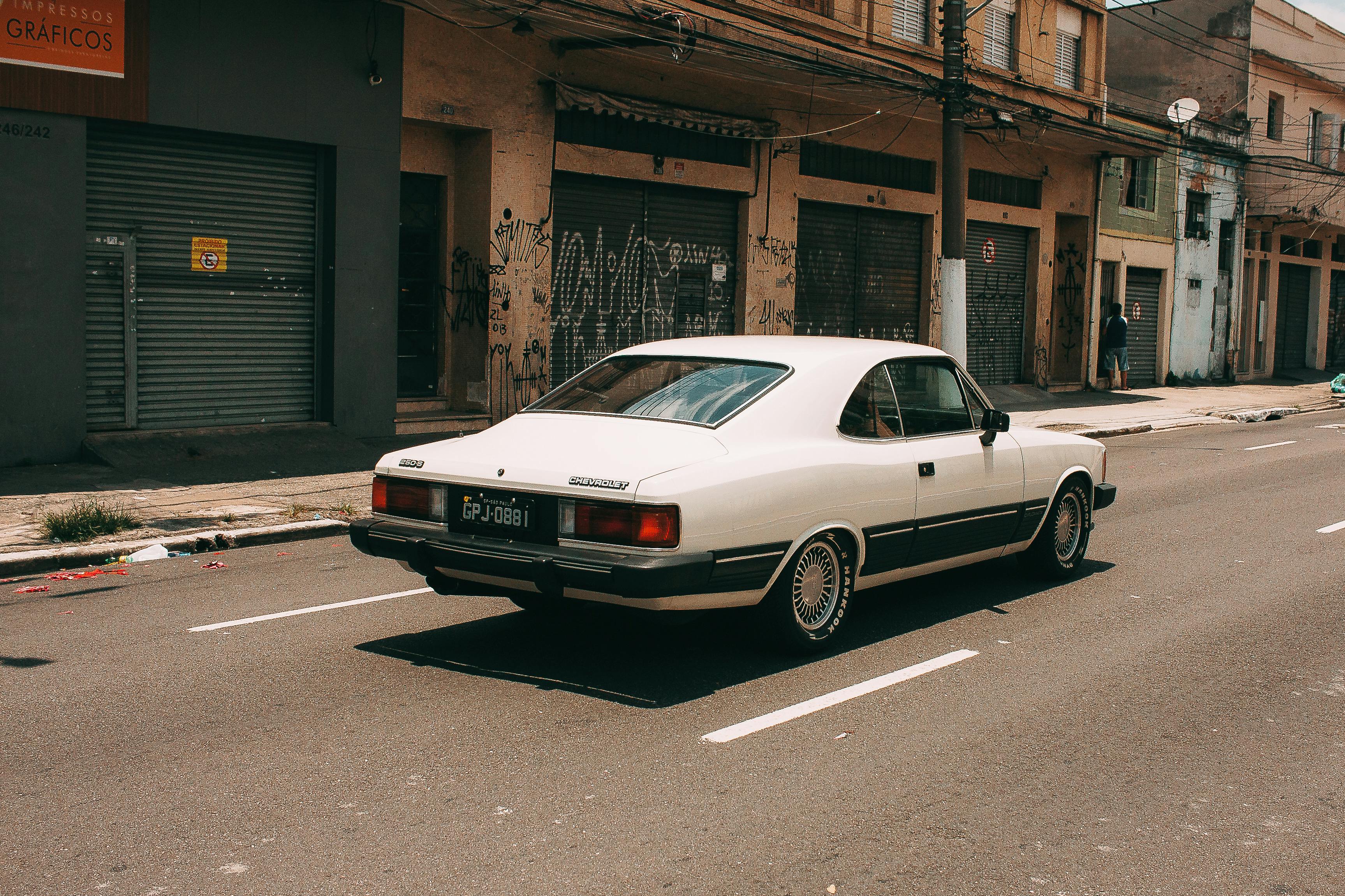 A vintage white Chevrolet Opala driving down an urban street lined with old storefronts.