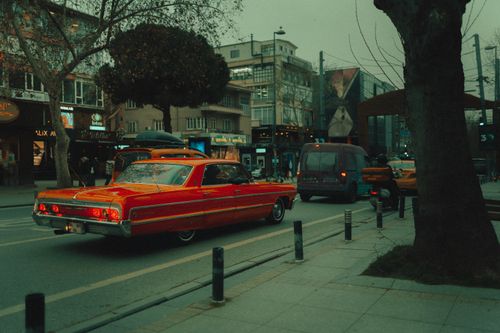 Free A classic red vintage car drives through a bustling city street lined with modern buildings and shops. Stock Photo