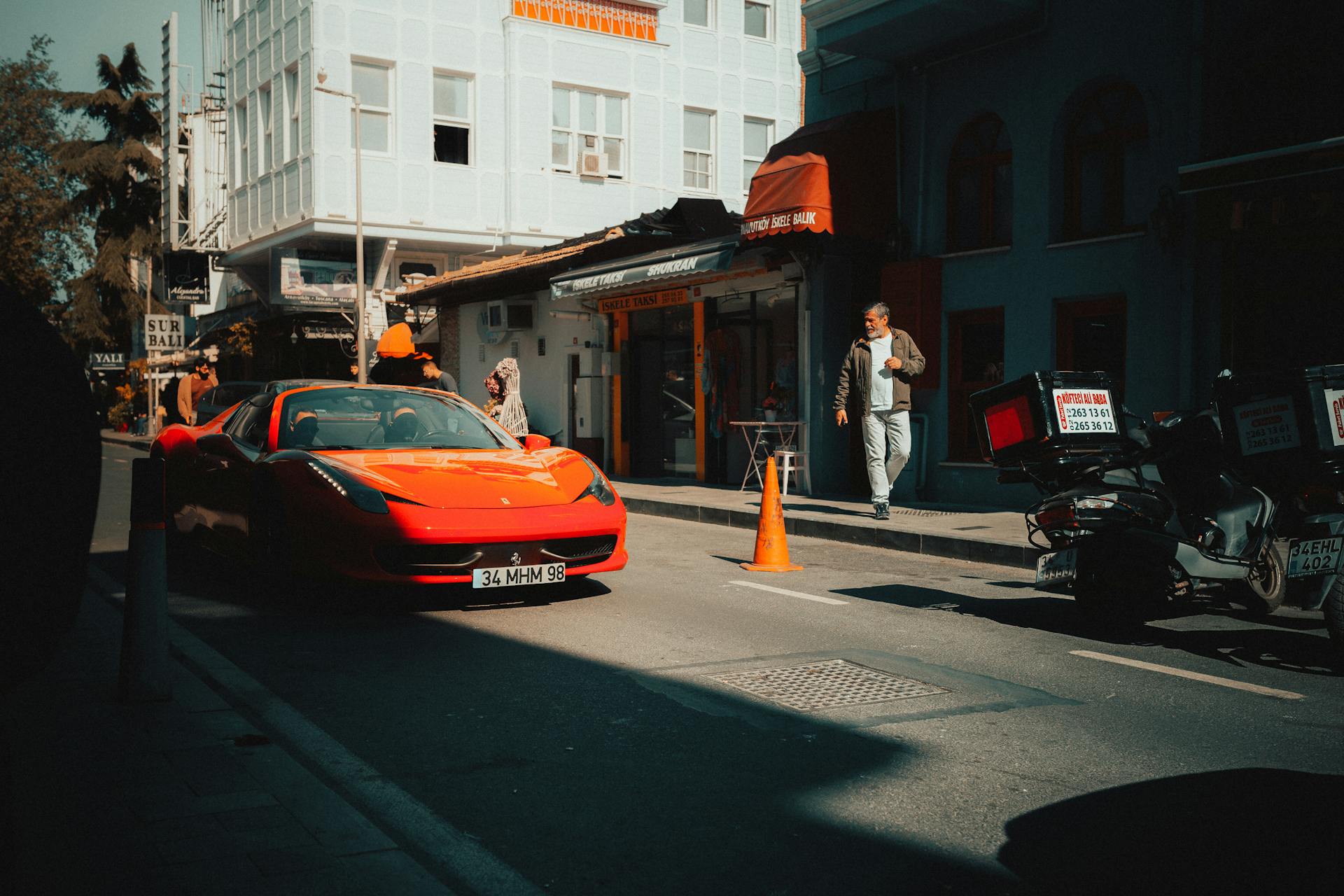 A striking orange Ferrari parked in a sunlit urban street setting with pedestrians and city life.