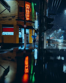 City street at night with neon hotel sign reflecting in a puddle, capturing urban ambiance.