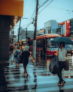 Pedestrians holding umbrellas cross a flooded street with buses during rainy day.
