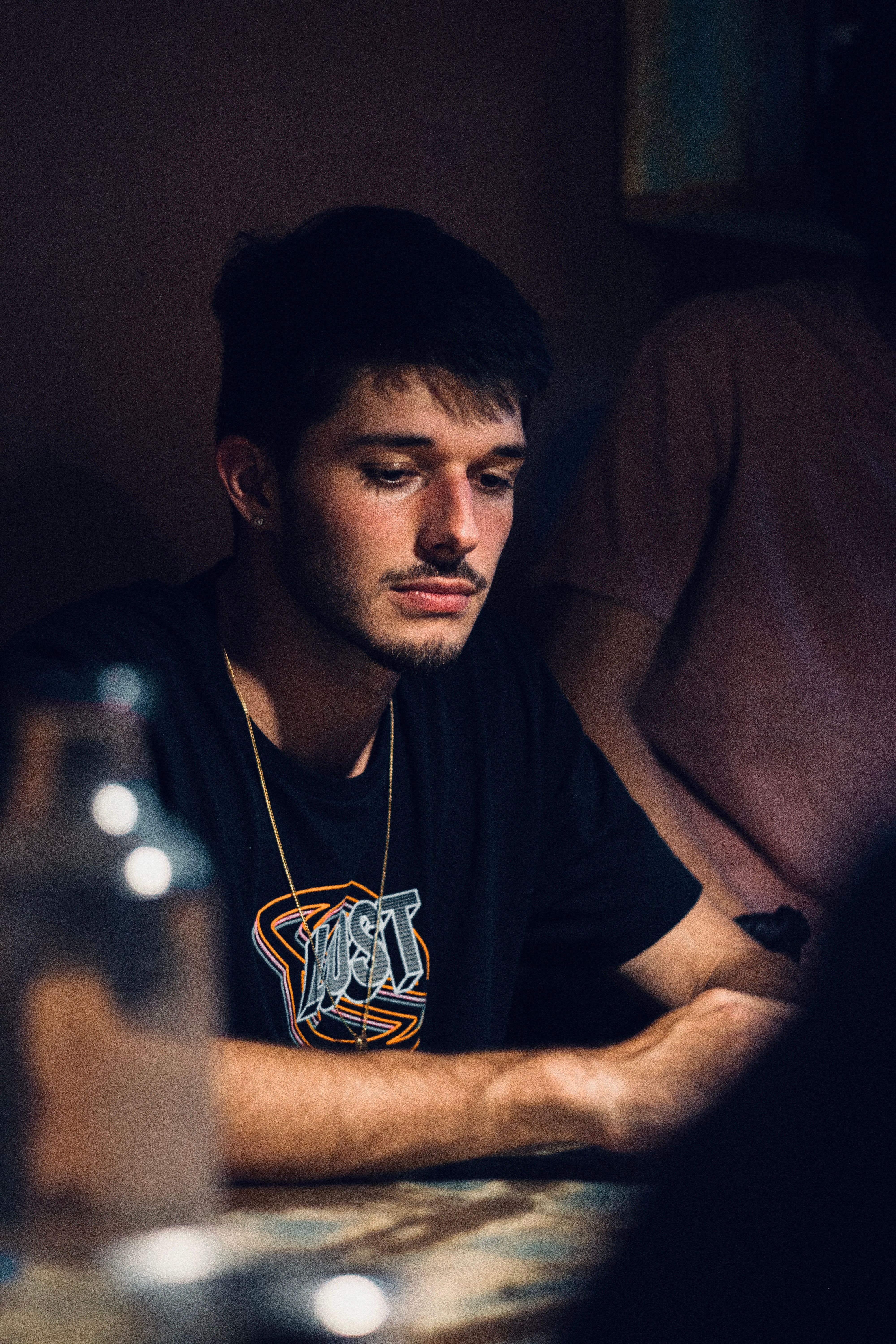 A young man in deep thought sits at a dimly lit table indoors.
