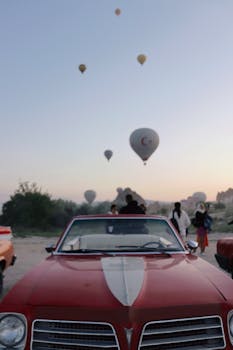 Front view of a classic red car with hot air balloons in the Cappadocia sky, at sunrise.