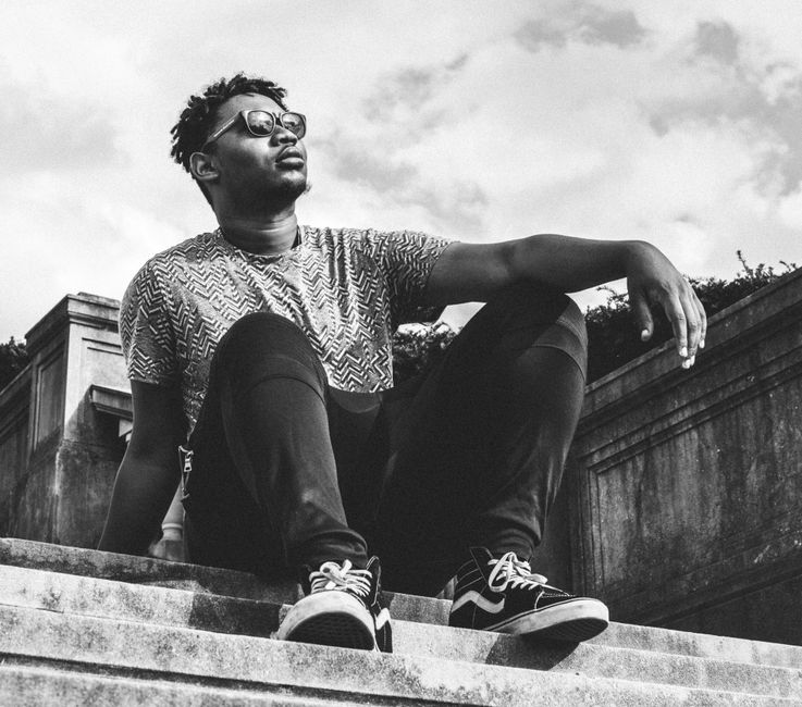 Black and white photo of a fashionable man sitting on steps outdoors, wearing sunglasses.